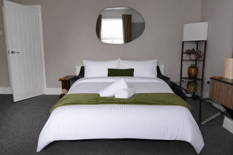 Bedroom with white bedding, green accent pillows, dark headboard, mirror, and shelving unit