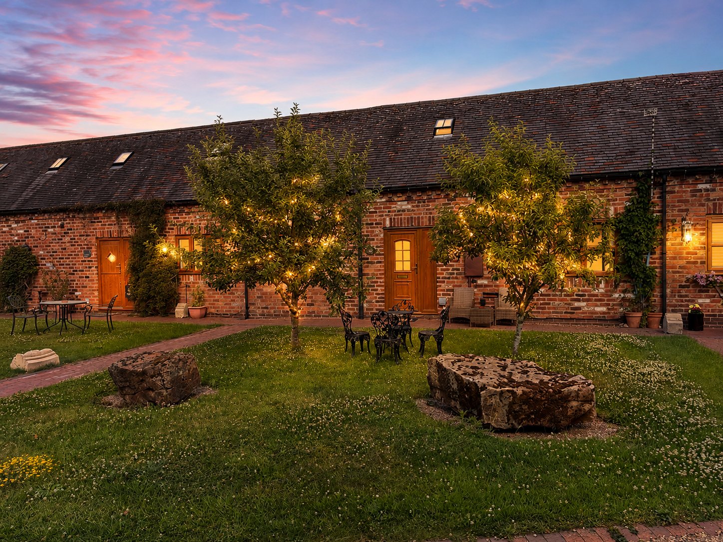 Brick cottage exterior at dusk with ivy-covered walls, illuminated windows, and manicured garden with seating area