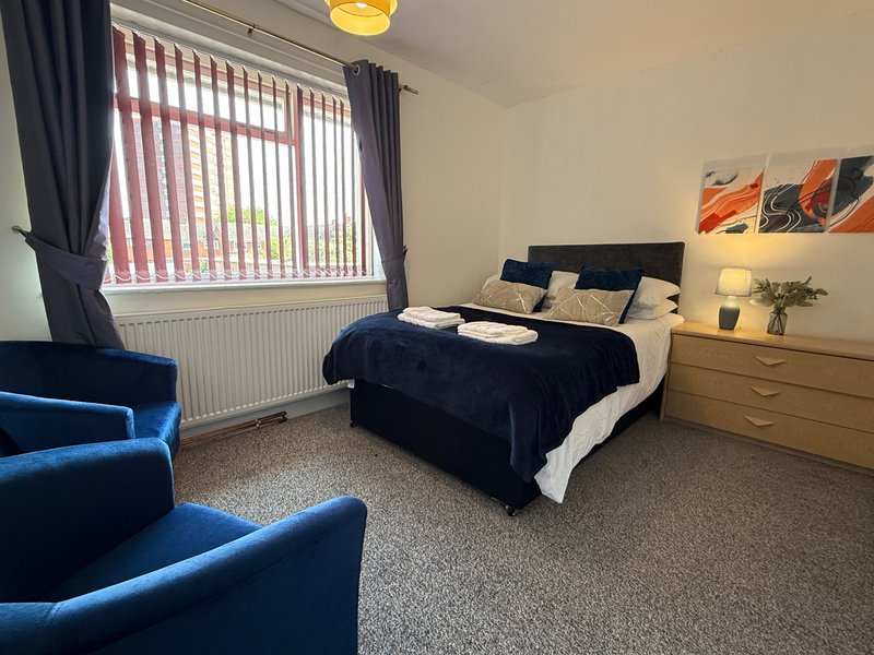 Spacious bedroom with double bed, blue armchairs, wooden dresser and vertical blinds
