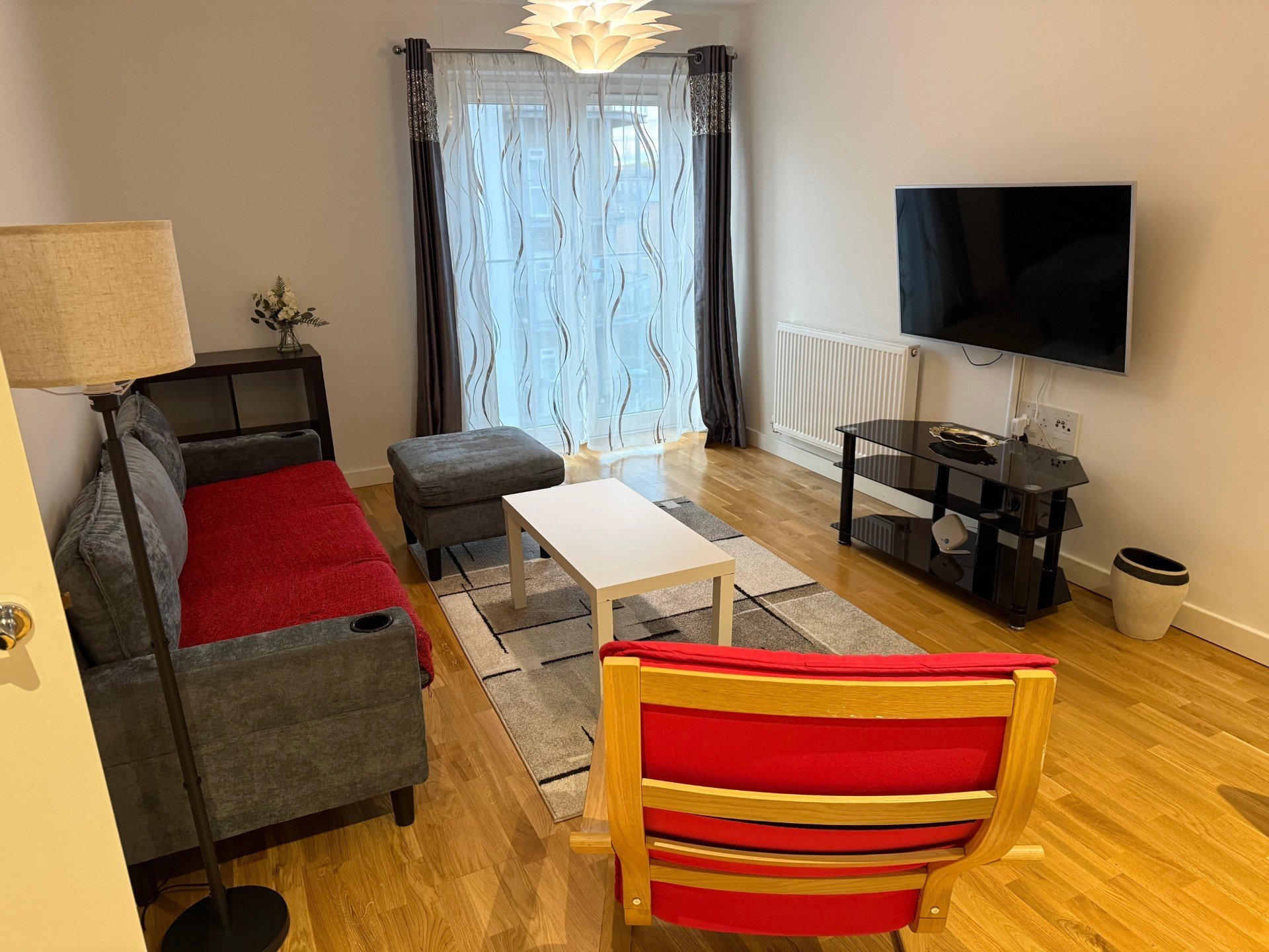 Living room with grey sofa, red accent chair, wooden floor, TV, and glass doors with patterned curtains