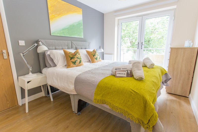 Bright bedroom with double bed, grey headboard, yellow throw, and garden view through patio doors