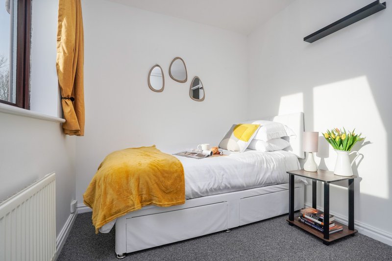 Bright modern bedroom with single bed, yellow throw, white bedding, side table with flowers and mirror wall decor