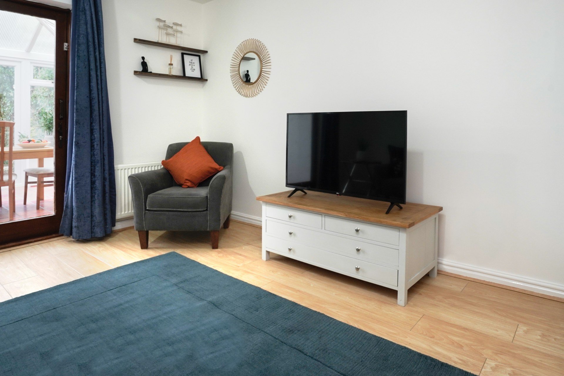 Living room with grey armchair, TV on wooden stand, blue rug and orange cushion