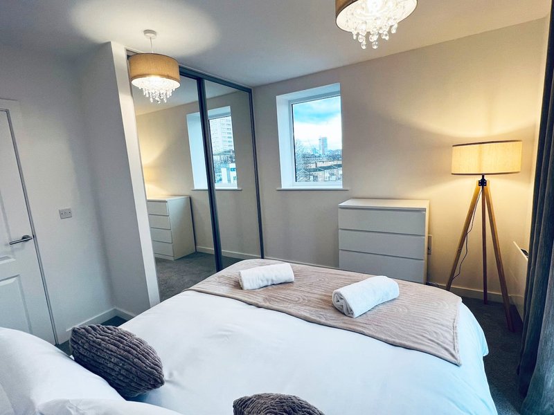 Modern bedroom with double bed, ensuite bathroom, floor lamp, and city views from windows