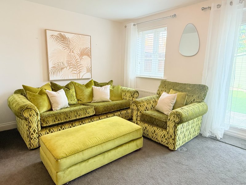 Living room with green upholstered sofa, armchair, yellow ottoman, and botanical wall art