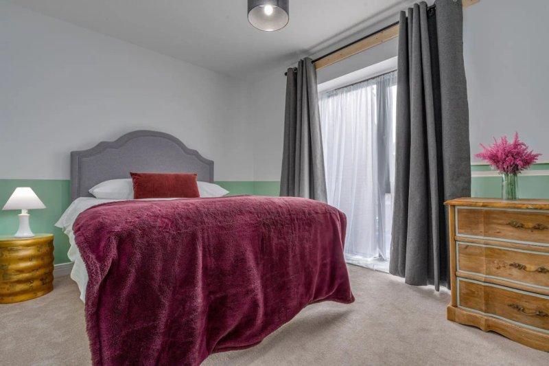 Bedroom with double bed, burgundy bedding, grey headboard, wooden dresser, and garden view