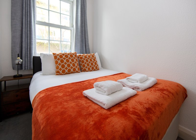 Bedroom with double bed, orange bedding, patterned cushions, white towels and window with grey curtains