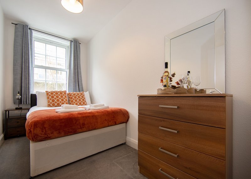 Bright bedroom with double bed, orange throw, wooden dresser with mirror and window with grey curtains