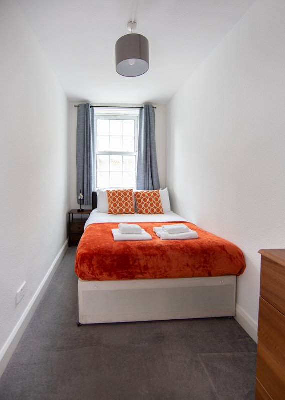 Compact bedroom with single bed, orange bedding, window with blue curtains, pendant light