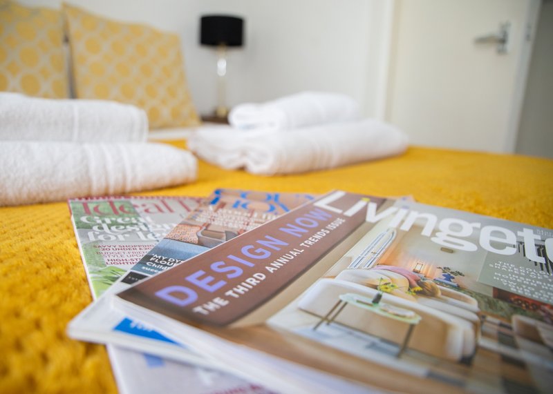 Bright yellow bedspread with white towels, magazines, and bedroom furnishings in background