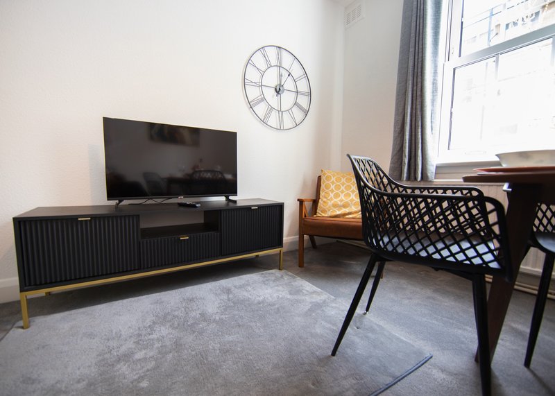 Modern living room with black TV unit, wall clock, grey rug, and black chair near window