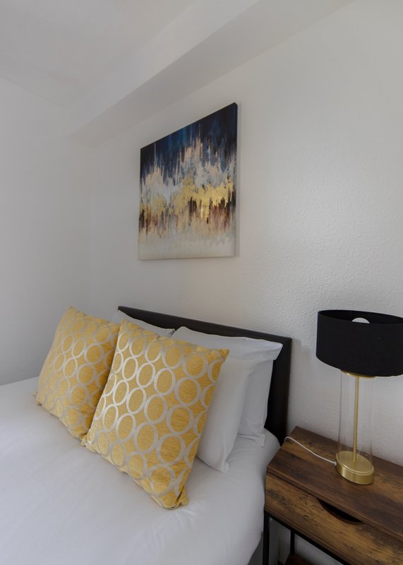 Bedroom with white bed, yellow patterned pillows, wooden bedside table, and abstract wall art