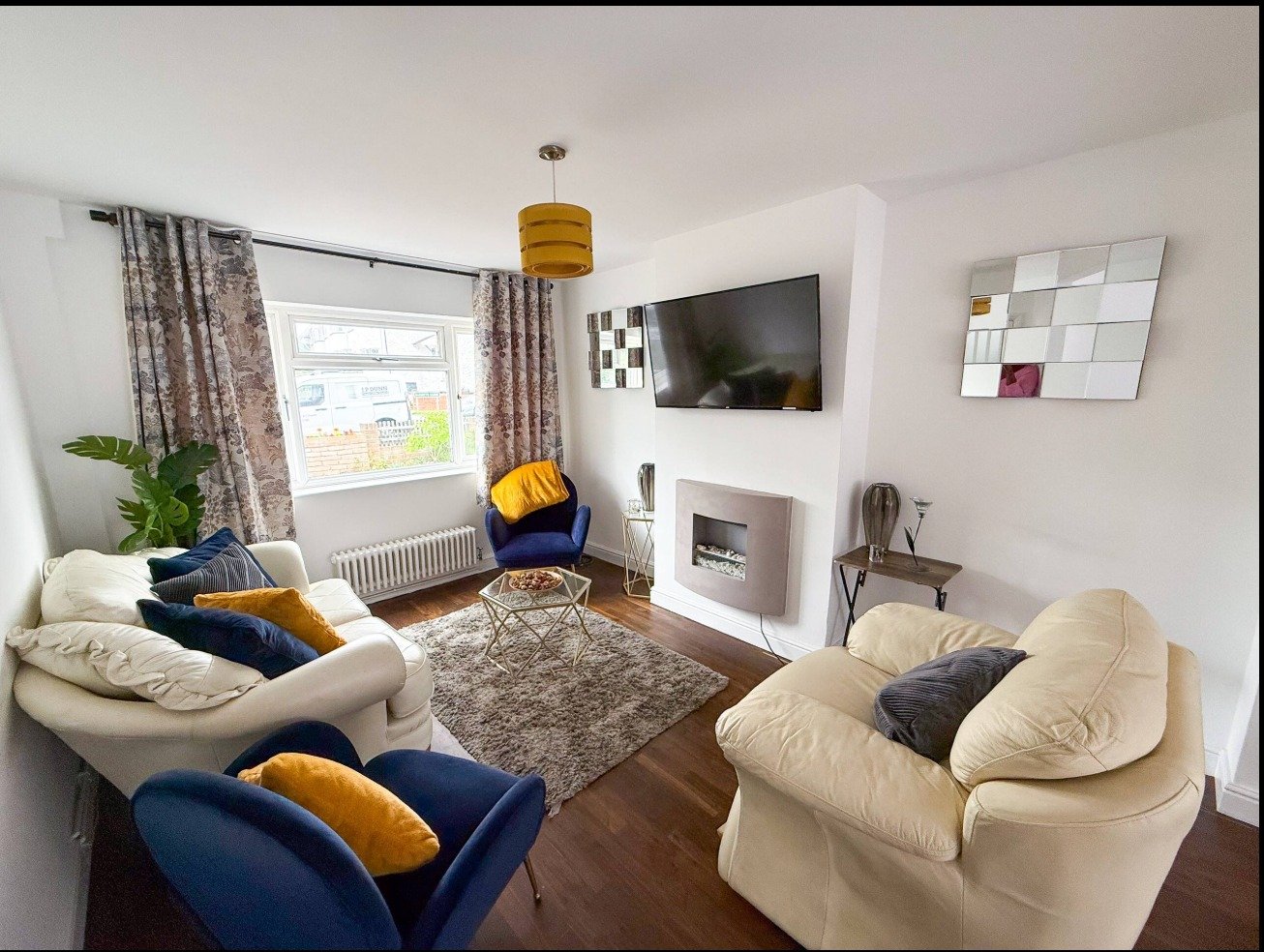 Bright living room with cream sofas, blue armchair, fireplace, wall-mounted TV, and large window