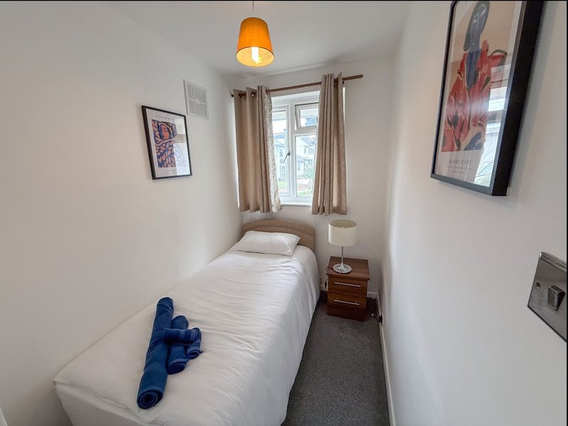 Compact bedroom with single bed, blue towels, wooden bedside table, window with curtains