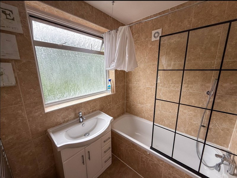 Bathroom with beige tile walls, curved sink, cabinet storage, bathtub with glass shower enclosure and window