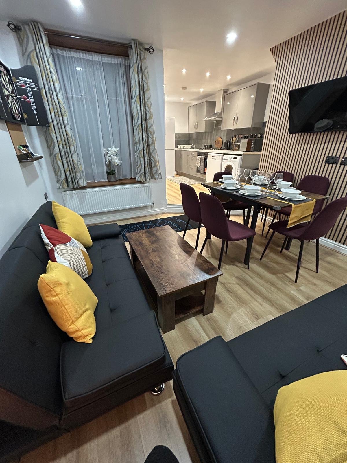 Modern open-plan living room with black sofa, yellow cushions, dining table and mounted TV