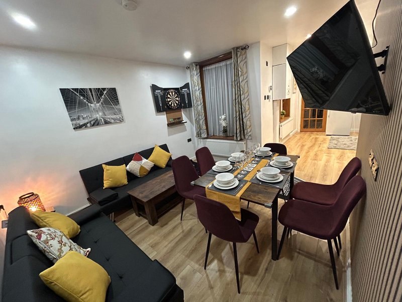 Open-plan living and dining area with black sofa, burgundy chairs, wooden dining table set, and wall-mounted TV