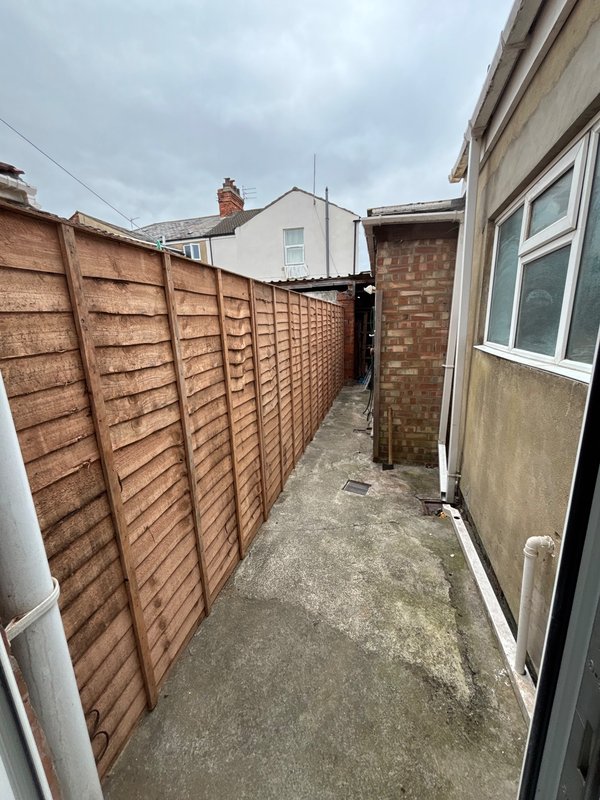 Narrow side passage with wooden fence, brick building wall, and concrete ground