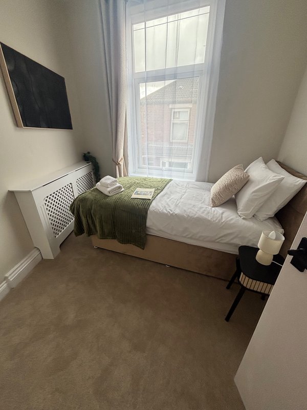 Bedroom with single bed, white pillows, green blanket, window with curtains, wall-mounted TV