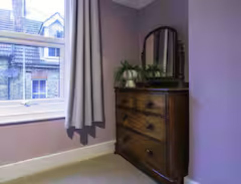 Bedroom corner with wooden dresser, arched mirror, potted plants, and window with curtains