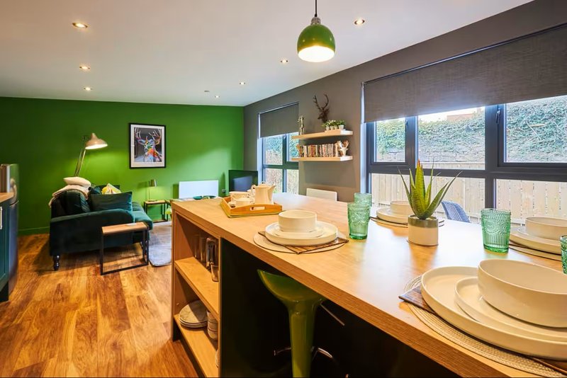 Open-plan dining and living room with wooden table, green accent wall and large windows overlooking hills