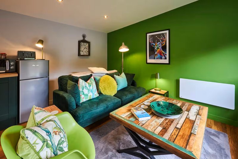 Modern studio with green accent wall, teal sofa, wooden coffee table and kitchenette