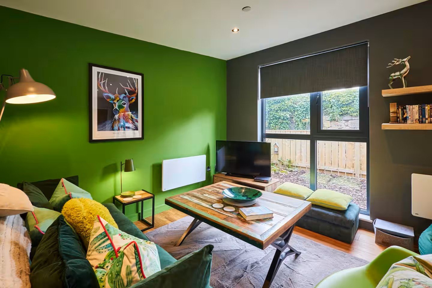 Modern living room with green accent wall, sofa, desk, and garden view through windows
