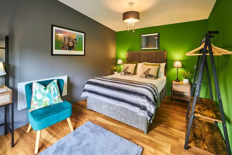 Bedroom with green accent wall, double bed, blue armchair, wooden floor and pendant lighting