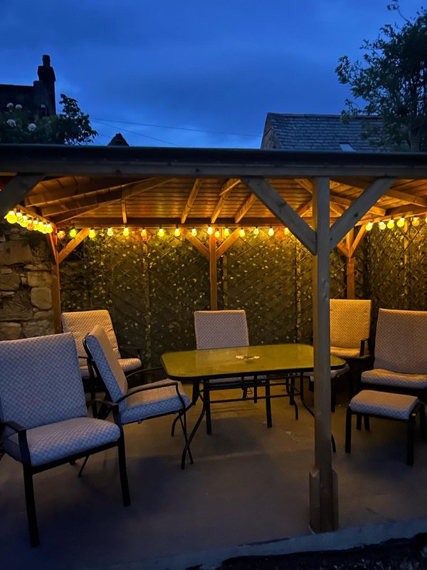 Covered patio at dusk with dining table, chairs, string lights, and house backdrop