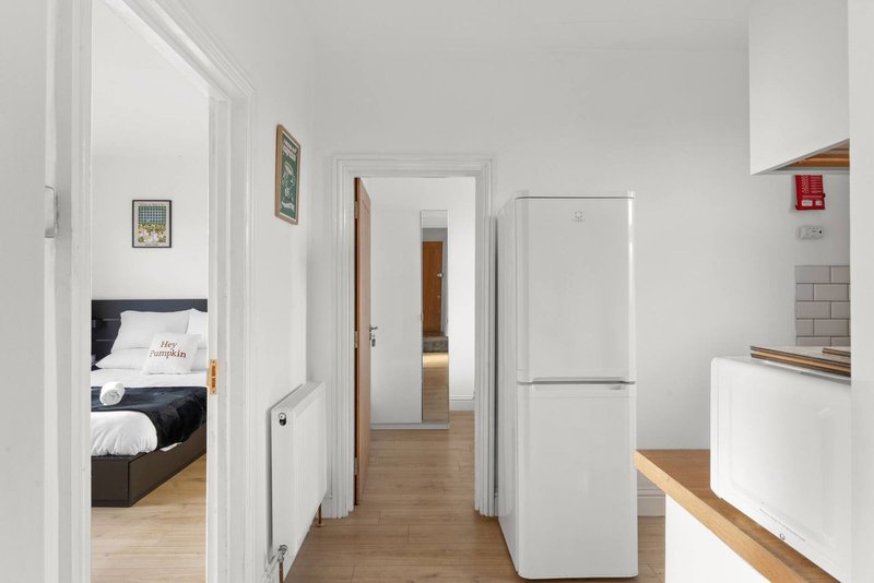 Bright hallway with white walls, wooden flooring, refrigerator and bedroom doorway visible