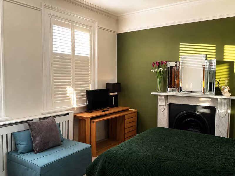Bedroom with green accent wall, double bed, wooden desk, TV, and fireplace mantelpiece