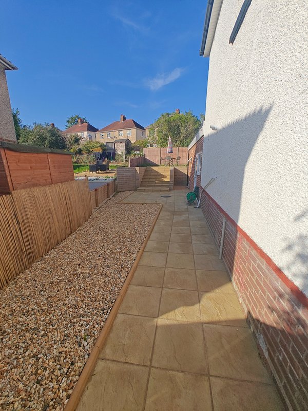 Narrow side pathway with paved surface, gravel border, wooden fence and brick walls, sunny day