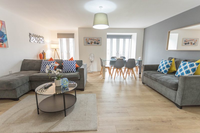 Open-plan living room with grey sofas, round coffee table, dining chairs and bright windows