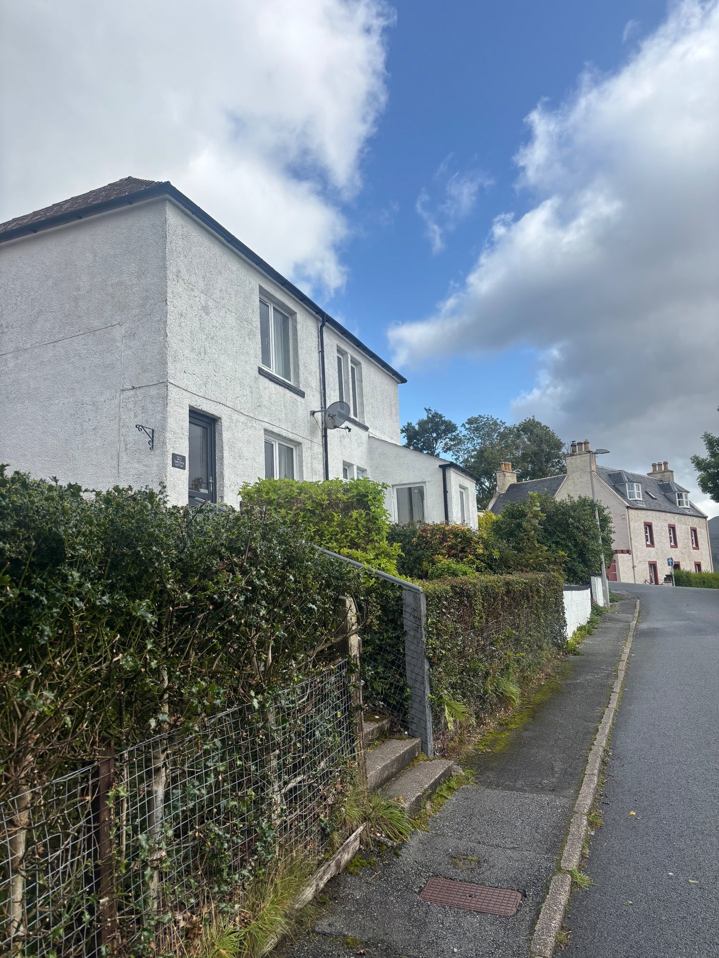 White rendered two-story house with dark roof, hedge fence, and street view