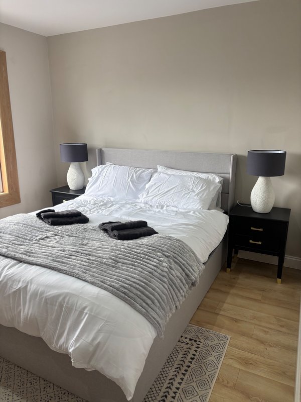 Bedroom with gray upholstered bed, white bedding, black nightstands, lamps, and wooden floor