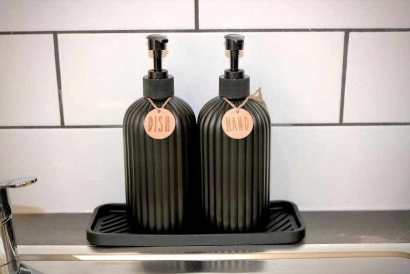 Two black ribbed soap dispensers with copper labels on bathroom counter with white subway tile backsplash