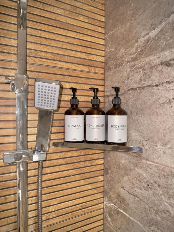 Bathroom shower area with wooden slat wall, three branded soap dispensers, and rainfall showerhead