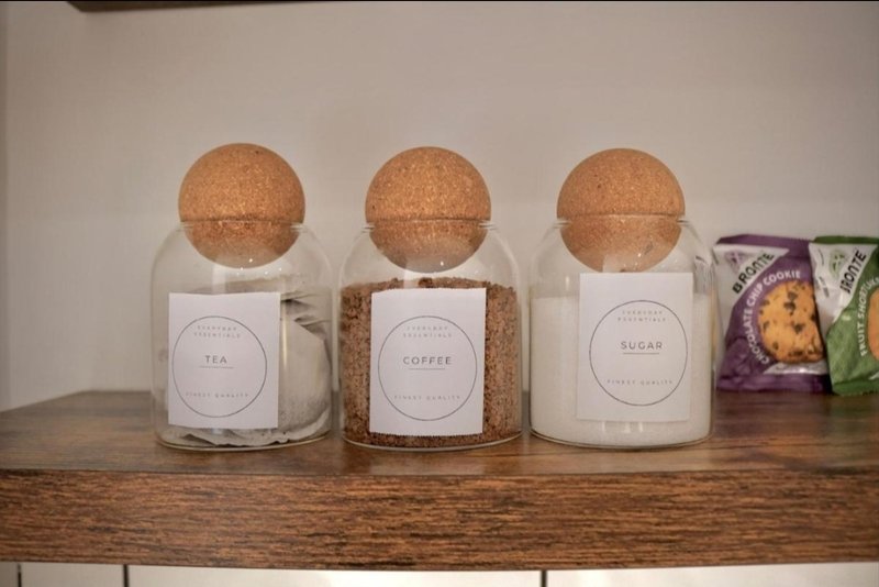 Three labeled kitchen storage jars with cork lids and packaged items on wooden shelf