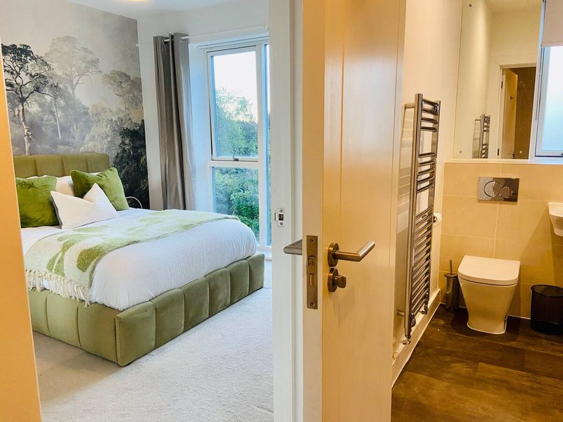 Modern bedroom with green upholstered bed, white bedding, landscape artwork, and ensuite bathroom visible
