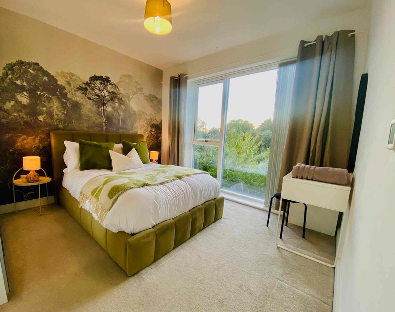 Modern bedroom with olive-green upholstered bed, white bedding, large landscape wall art, and floor-to-ceiling windows with garden views