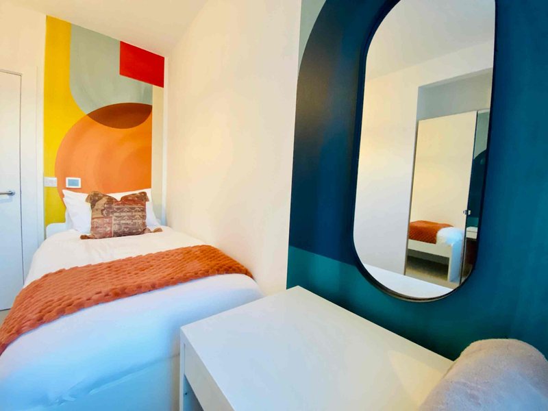 Compact bedroom with single bed, colorful geometric headboard art, and oval mirror on teal wall
