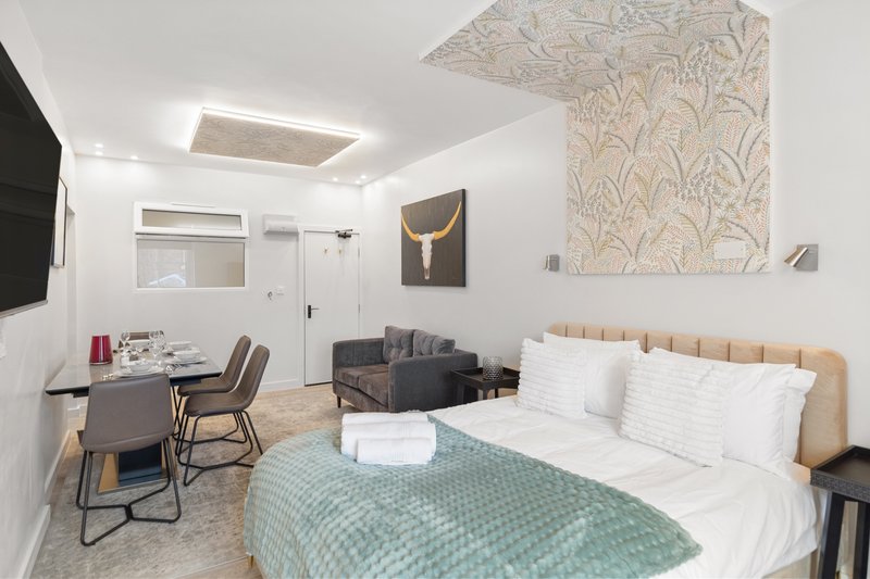 Modern studio bedroom with double bed, desk area, seating, and kitchenette in neutral tones