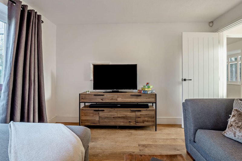 Living room with TV on wooden stand, gray sofa, light armchair, and curtained window
