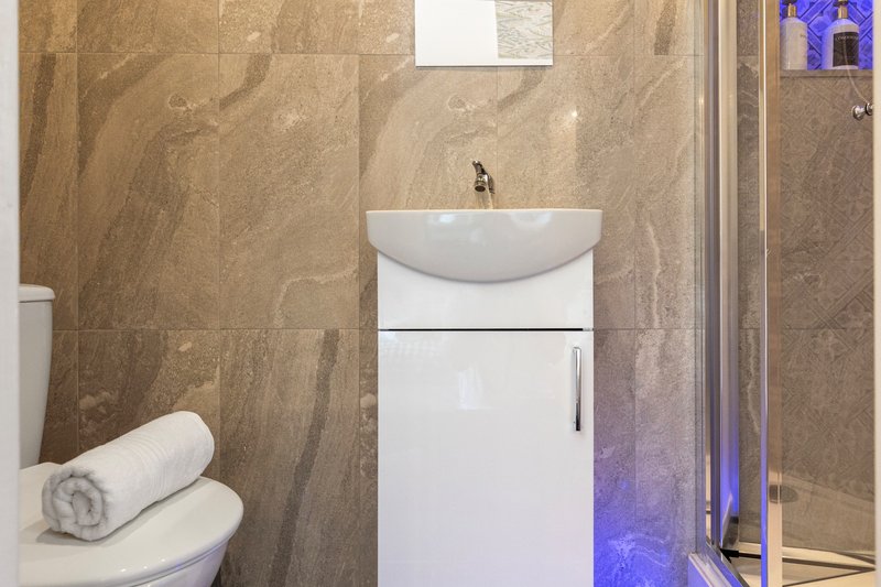 Modern bathroom with beige tile walls, white pedestal sink, rolled towel, and glass shower enclosure