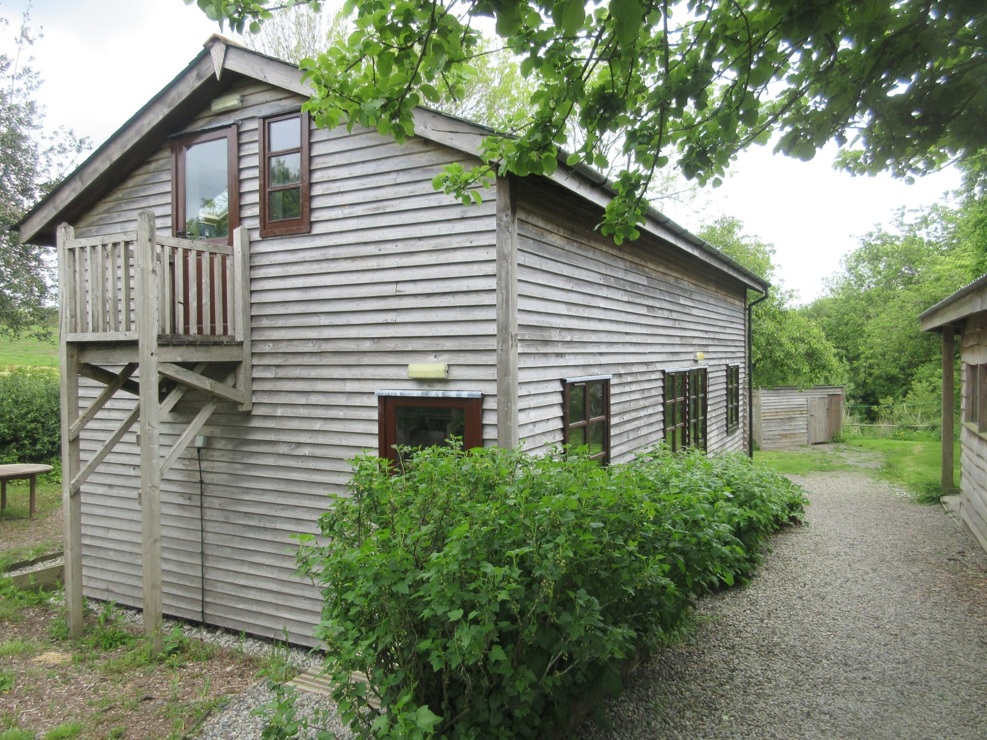 Modern wooden lodge exterior with elevated deck, surrounded by green landscaping and gravel path.