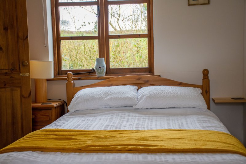 Bedroom with wooden bed, white pillows, yellow throw blanket, and large windows overlooking countryside.