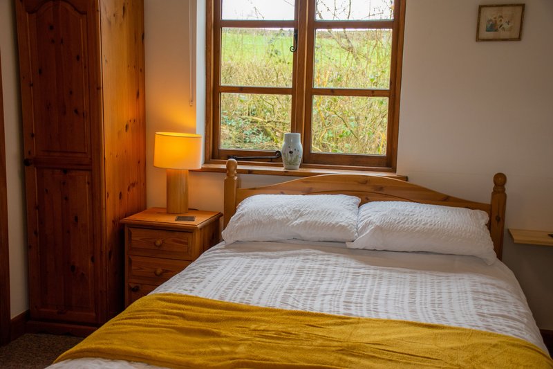 Bedroom with double bed, wooden wardrobe, bedside table with lamp and windows overlooking garden
