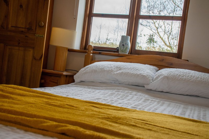 Bright bedroom with wooden bed frame, white pillows, yellow throw blanket, and large windows overlooking bare trees.