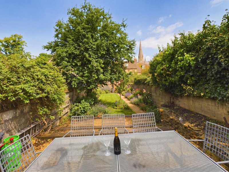 Garden view from terrace with table, mature trees, stone walls and church spire beyond