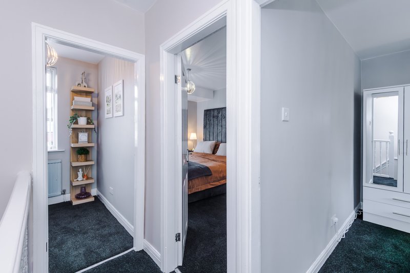 Hallway with open doorways leading to bedroom and storage areas, white walls and dark carpet